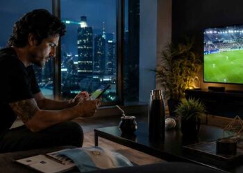 Hombre revisando apuestas en línea mientras ve un partido de fútbol en televisión